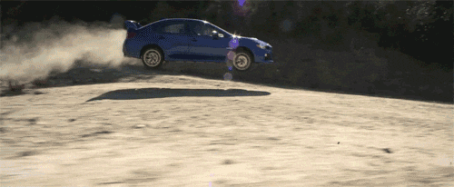 A side view of a blue car speeding along a dirt road. As it drives down the hill it catches some air, and lands with dust trailing behind it.