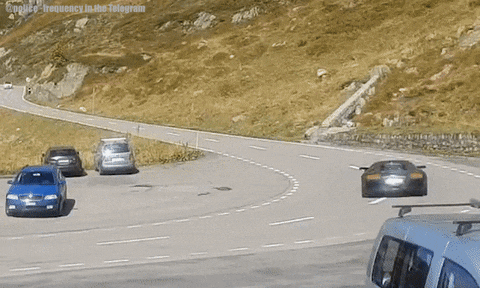 A car drives up a winding road, past parked vehicles, loses control and spins onto the hillside shoulder and flips over.