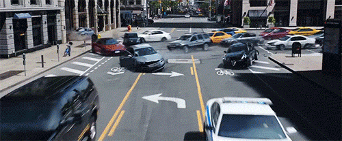 A large crowd of vehicles speed around a city block corner, some crashing into one another and others speeding along.
